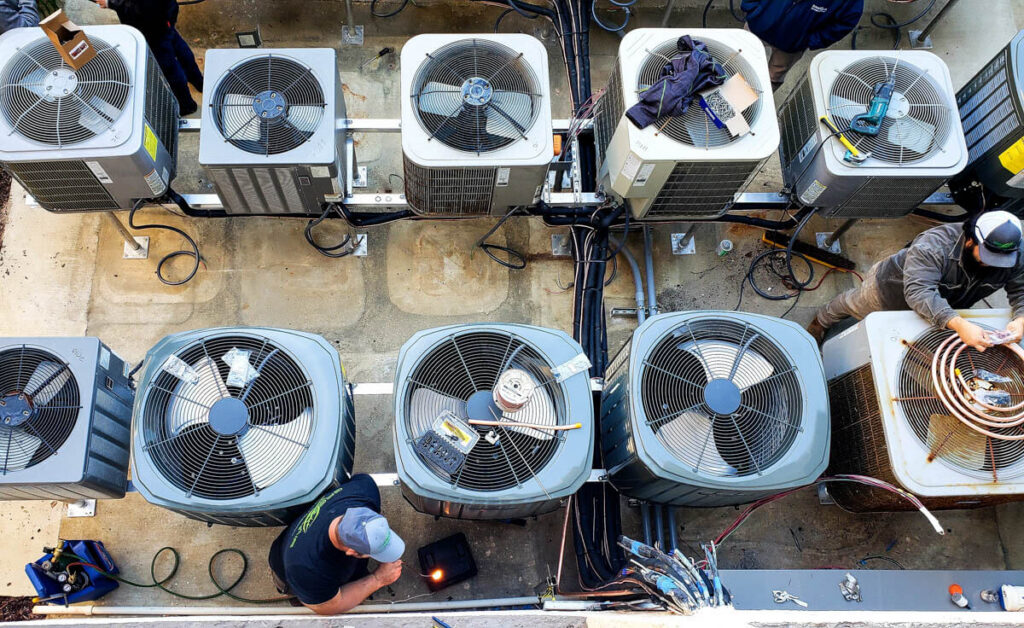 Aerial view of HVAC technicians working on multiple outdoor air conditioning units, with tools and equipment laid out on top of and around the units.