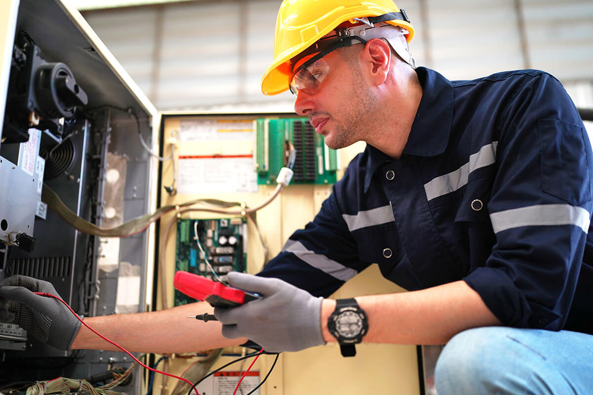 A technician wearing a yellow hard hat, safety glasses, and gloves uses a multimeter to test HVAC electrical components inside an open industrial control panel.